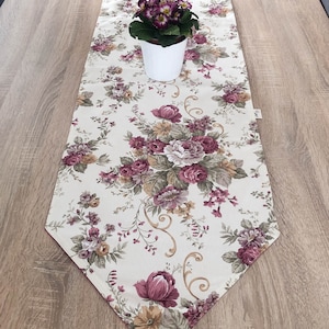Op de afbeelding: Een witte tafelrunner met een bloemenpatroon met roze en bruine rozen, groene bladeren en gouden accenten. De runner ligt op een houten tafel met een geplante plant met paarse bloemen.