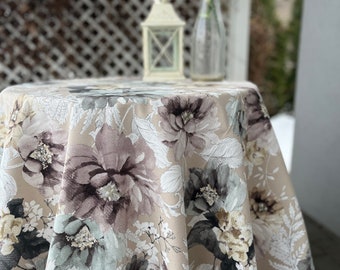 May include: A beige round tablecloth with a floral pattern featuring large, watercolor-style flowers in shades of brown, gray, and white. The tablecloth is draped over a table and has a slightly ruffled edge.