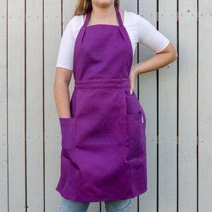 Puede incluir: Un delantal morado con dos bolsillos grandes. El delantal está hecho de una tela gruesa y duradera y tiene un diseño simple y clásico.