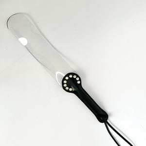 Puede incluir: Una paleta de plástico transparente con un mango de cuero negro y tachuelas plateadas. El mango tiene un lazo para colgar.