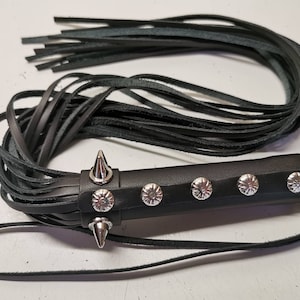 Puede incluir: Látigo de cuero negro con mango trenzado y tachuelas plateadas. El látigo tiene una larga franja y un lazo para colgar.