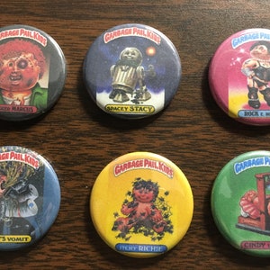May include: Six round, colorful buttons with images of cartoon characters from the Garbage Pail Kids trading card series. The characters are:  Mugg'd Marcus, Spacey Stacy, Rock E. Horror, Haley's Vomit, Itchy Richie, and Cindy Lopper.