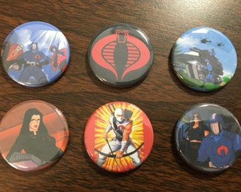 Könnte beinhalten: Sechs runde Anstecknadeln mit Figuren und Symbolen aus der G.I. Joe Zeichentrickserie. Die Nadeln zeigen ein rotes Kobra-Logo, eine blaue Figur mit rotem Helm, eine weibliche Figur mit schwarzen Haaren und einem roten Kobra-Logo, eine blaue Figur mit rotem Helm und einem roten Kobra-Logo, eine blaue Figur mit rotem Helm und einem roten Kobra-Logo sowie eine weiße Figur mit rotem Helm und einem roten Kobra-Logo.