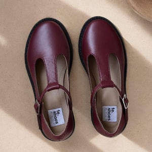 May include: A pair of burgundy leather T-strap shoes with black soles. The shoes have a buckle closure and the brand name "te. shoes KYIV" is visible on the insole.
