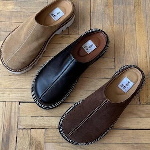 May include: Three pairs of slip-on shoes in tan suede, black leather, and brown suede. Each shoe has a rounded toe, a thick sole, and a stitched design around the edges. The shoes are displayed on a wooden floor.