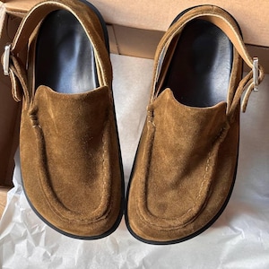May include: A pair of brown suede shoes with a strap and buckle fastening. The shoes have a rounded toe and a black sole. The shoes are on white paper inside a cardboard box.