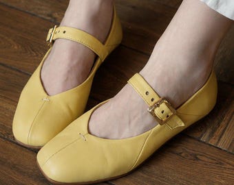 May include: A pair of bright yellow leather Mary Jane flats featuring a square toe, a single strap across the instep, and a gold-toned metal buckle closure. The shoes have a smooth finish and a low-profile design with visible stitching at the toe.