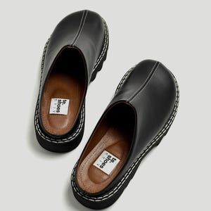 May include: A pair of black leather clogs with a thick, white-stitched sole. The clogs have a rounded toe and a slip-on design. The interior features a brown insole with white text. The shoes are displayed on a light background.