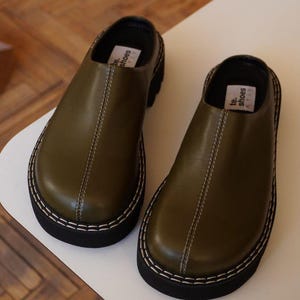 Puede incluir: Un par de zuecos de cuero verde oliva con suela negra y costuras blancas. Los zuecos tienen una puntera redondeada y un tacón bajo. La marca "te. shoes" está impresa en una pequeña etiqueta en el interior.