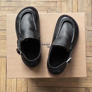 Può includere: Un paio di zoccoli in pelle nera con chiusura a fibbia, appoggiati su una scatola di cartone. Gli zoccoli hanno la punta arrotondata e una fodera in montone. La scatola riporta la scritta "SHOES KYIV".