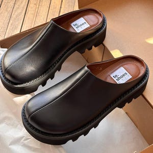Op de afbeelding: Een paar zwarte leren klompen met een dikke, gegroefde zool. De klompen hebben een ronde neus en een glad bovenwerk. De binnenkant is bruin met een label "te. shoes KYIV". De klompen zitten in een kartonnen doos.