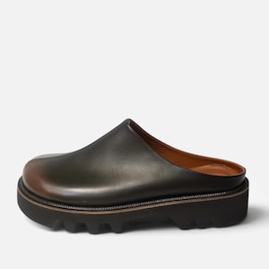 May include: A dark olive green leather mule with a smooth, rounded toe and a prominent center seam. The shoe features a thick, black lugged rubber platform sole with a visible welt and a tan leather interior lining, showcasing a slip-on design.