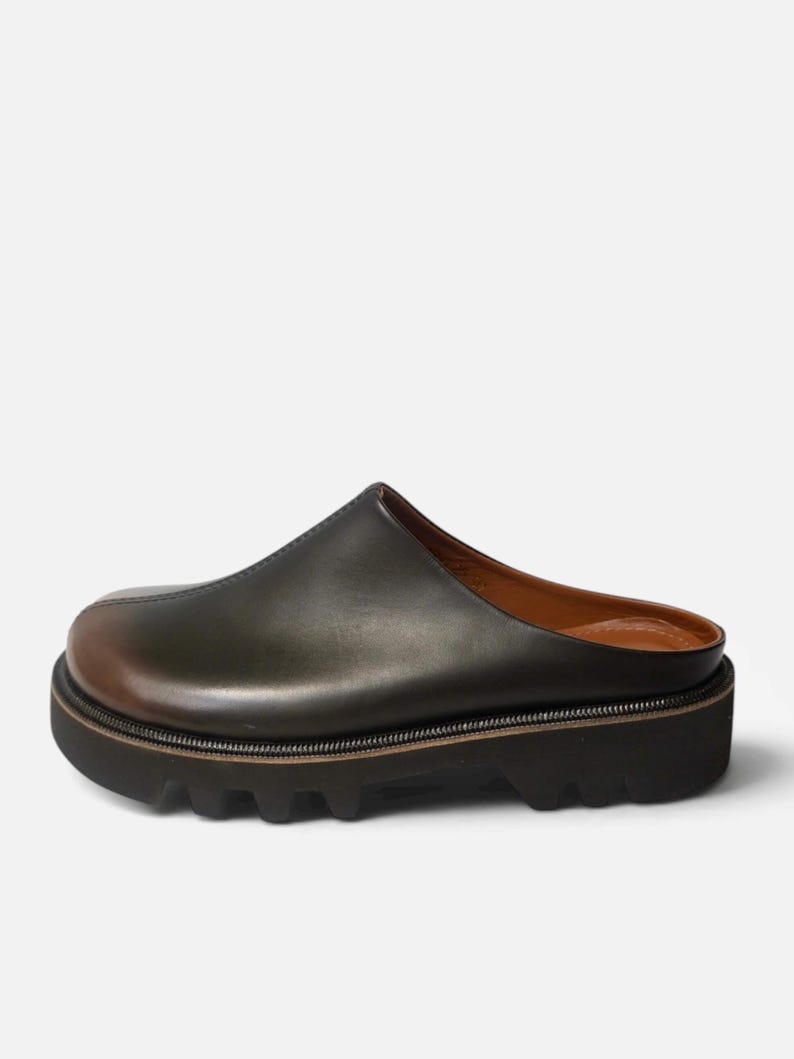 May include: A dark olive green leather mule with a smooth, rounded toe and a prominent center seam. The shoe features a thick, black lugged rubber platform sole with a visible welt and a tan leather interior lining, showcasing a slip-on design.