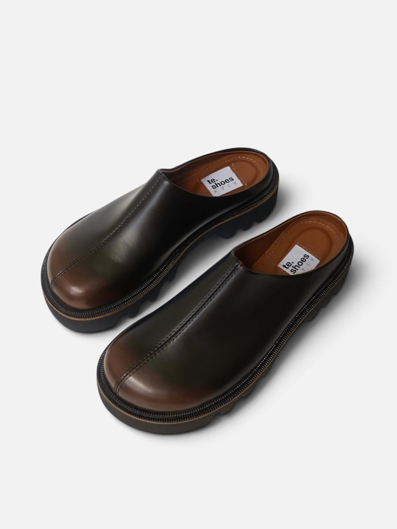 May include: A pair of dark brown leather slip-on mules with a rounded toe, a thick black lug sole, and a visible center seam. The interior footbed features a white label with the black text te. shoes KYIV printed on the heel area of each shoe.