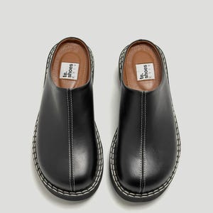 Puede incluir: Par de zuecos de cuero negro con costuras blancas y suela gruesa. Los zuecos tienen una puntera redondeada y un diseño sin cordones. El nombre de la marca "te. shoes KYIV" está impreso en la plantilla.