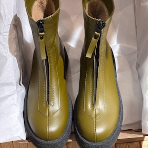 May include: Olive green ankle boots with a black zip and a thick black sole. The boots have a rounded toe and a soft, light brown interior. The boots are in a cardboard box with white tissue paper.