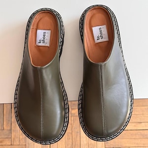 May include: A pair of olive green leather clogs with a rounded toe and a thick black sole. The clogs have a stitched white border and a tan insole. The text "te. shoes KYIV" is printed on a white label inside each shoe.