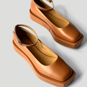 May include: A pair of tan leather flats featuring a square toe, a thick platform sole, and a thin ankle strap with a metal buckle. The interior of the shoe displays the text 248 0388/24 printed in black on the light tan insole.