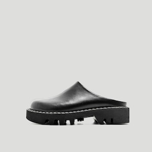 Pode incluir: Tamanco de couro preto com sola grossa e com ranhuras. O sapato tem bico arredondado e salto baixo. A sola tem aproximadamente 5 cm de espessura. A parte superior é lisa e as costuras são visíveis.