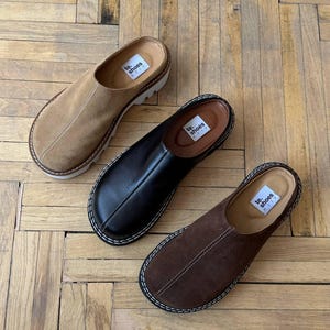 Pode incluir: Três pares de sapatos slip-on em cores diferentes: camurça bege, couro preto e camurça marrom. Cada sapato tem uma ponta arredondada, uma sola grossa e costuras visíveis. Os sapatos são exibidos em um piso de madeira.