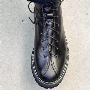 Könnte beinhalten: Ein schwarzer Schnürstiefel aus Leder mit abgerundeter Zehenpartie, einer markanten Naht entlang der Mitte des Vorderblatts und einer dicken schwarzen Profilsohle mit auffälligen weißen Kontrastnähten entlang des Schuhrahmens.