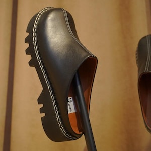 May include: Black leather clogs with a chunky, lugged sole and white stitching. The shoes have a slip-on design and a rounded toe. The interior is a light brown color. The word "shoes" is visible on the side of the shoe.