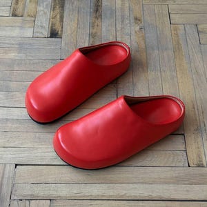 Könnte beinhalten: Ein Paar leuchtend rote Lederclogs. Die Clogs haben eine runde Spitze und einen niedrigen Absatz. Die Schuhe stehen auf einem Holzboden.