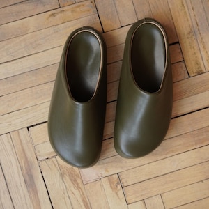May include: A pair of olive green leather clogs with a rounded toe and a gold-colored trim. The clogs are placed on a wooden parquet floor. The shoes have a simple, minimalist design.