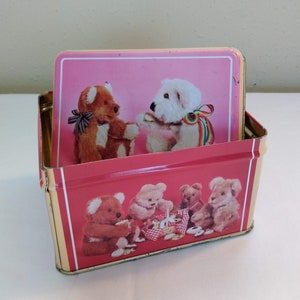 May include: A pink metal tin with a picture of teddy bears on the front. The lid has a picture of two teddy bears, one brown and one white, holding hands. The tin is about 5 inches wide and 3 inches tall.