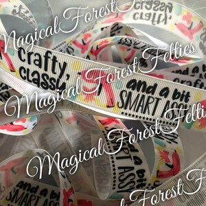 May include: White ribbon with black text that reads "crafty, classy, and a bit SMART ASSY". The ribbon is folded and layered, showing the text multiple times. The ribbon is from Magical Forest Felties.