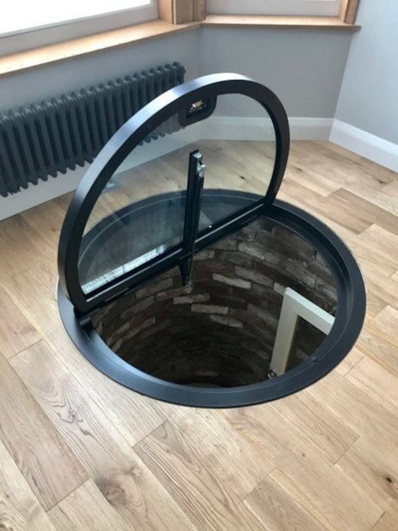 May include: A black circular trapdoor with a glass panel and a metal frame. The trapdoor is open and reveals a brick-lined shaft leading down into a basement.