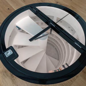 May include: A circular, black-framed door with a glass panel opens to reveal a spiral staircase leading down into a white wine cellar.