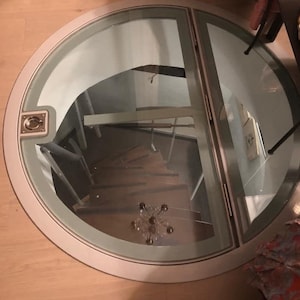 May include: A circular, glass-covered opening in a wooden floor, revealing a spiral staircase leading down. The opening has a silver frame and a handle.