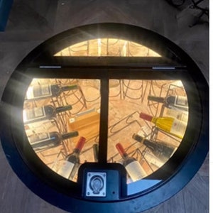 May include: A round, black, metal door with a glass panel revealing a wine cellar below. The cellar is lit with white lights and contains rows of wine bottles.