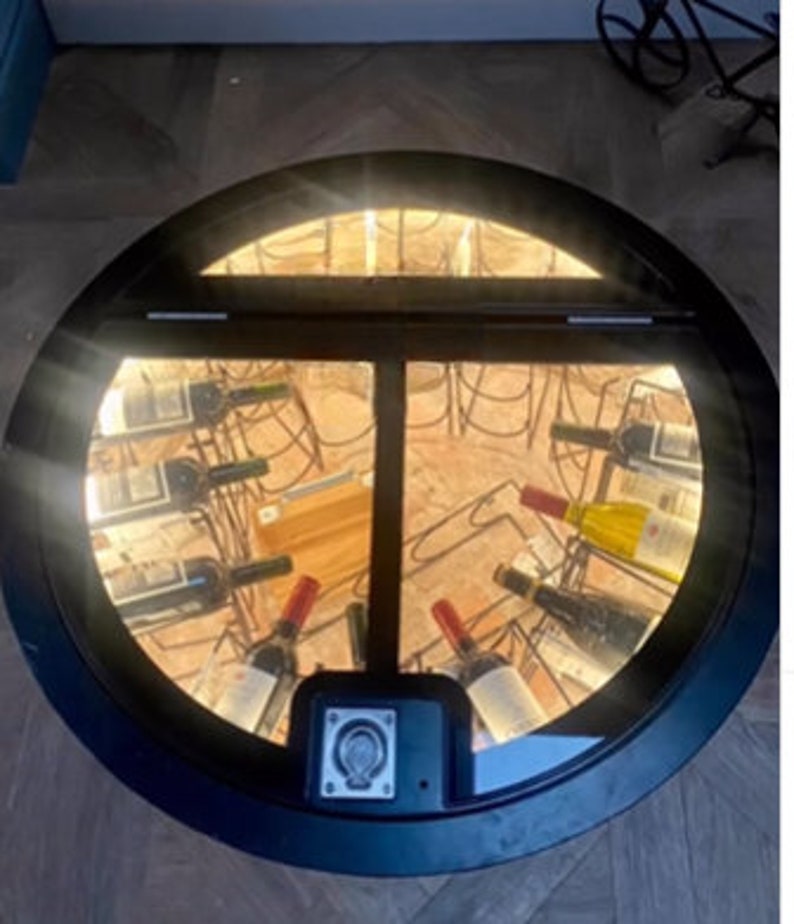 May include: A round, black, metal door with a glass panel revealing a wine cellar below. The cellar is lit with white lights and contains rows of wine bottles.