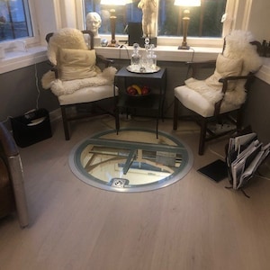 May include: A round, glass trapdoor in a light wood floor. Two white chairs with sheepskin throws are positioned on either side of the trapdoor. A small black table with a bowl of fruit sits between the chairs. Two lamps with white shades are on the windowsill behind the chairs.