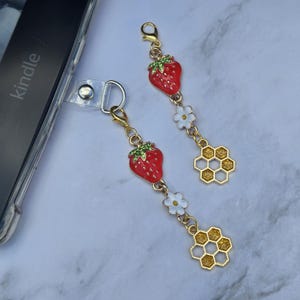 May include: Two gold-colored phone charms with red strawberry pendants, white flower accents, and honeycomb charms. The charms are attached to a clear phone case with the word "kindle" visible.