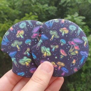May include: Two round black coasters with a rainbow mushroom pattern on a dark background. The mushrooms are outlined in white and have a glittery effect.