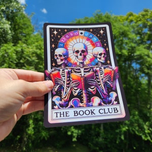 May include: A tarot card with a black background and a colorful illustration of three skeletons reading books. The card has the text "The Book Club" at the bottom.