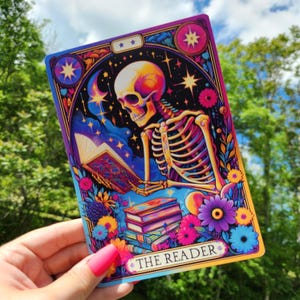 May include: A tarot card with a colorful illustration of a skeleton reading a book. The card is titled "The Reader" and has a black background with stars and a crescent moon. The skeleton is wearing a blue robe and is sitting on a pile of books. The card is surrounded by flowers.