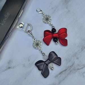 May include: Two decorative charms with lobster clasps. One charm features a red bow, black beads, and a silver spiderweb pendant. The other charm has a black bow, white beads, and a silver spiderweb pendant. The charms are designed to attach to a device.