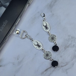 May include: Two silver-toned phone charms with a Halloween theme. Each charm features an oval pendant with a black spider design, a spiderweb charm, and a black bead. The charms are attached to a clear phone strap.