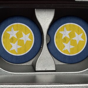 May include: Two round coasters with a blue background and a yellow circle in the center. The circle has three white stars inside. The coasters are in a car cup holder.