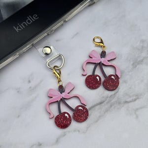 May include: Cherry-shaped phone charms with pink bows and glittery red cherries. One charm is attached to a clear phone strap, while the other is separate. The charms have gold-colored clasps and are set against a marble background.