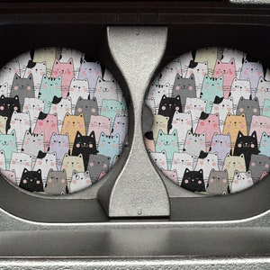 May include: A set of two round, gray coasters with a colorful pattern of cartoon cats. The cats are in various poses and colors, including pink, blue, yellow, and gray.