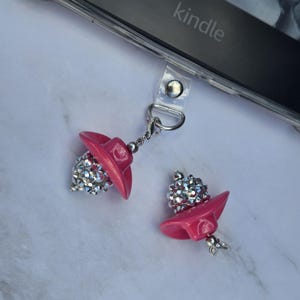 May include: Two pink cowboy hat charms with silver rhinestone accents. One charm is attached to a clear phone case with a silver chain. The word "kindle" is visible on the phone case.