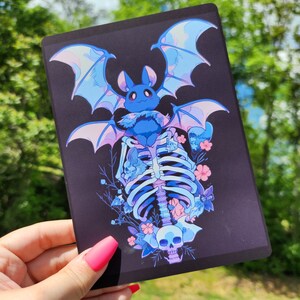 May include: A dark rectangular print featuring a blue bat with large wings, perched atop a skeleton. The artwork includes blue and pink floral accents and a skull at the bottom, all set against a black background.
