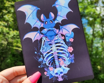 Puede incluir: Una impresión rectangular oscura con un murciélago azul con grandes alas, posado sobre un esqueleto. La obra de arte incluye detalles florales azules y rosas y una calavera en la parte inferior, todo sobre un fondo negro.