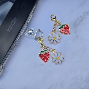 May include: Two gold-colored phone charms with red strawberry and white daisy flower pendants. The charms are attached to a clear phone case with the word "kindle" visible on the device.
