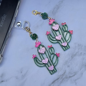 May include: Two green glitter cactus charms with pink heart accents and gold-colored hardware. Each cactus has three glittery pink heart details and pink flower accents. The charms are attached to a clear phone case.